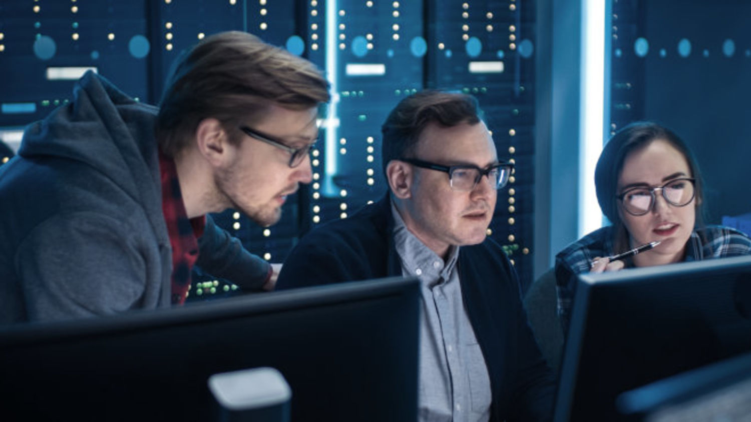 Two men and a woman sit in front of several monitors in a data centre